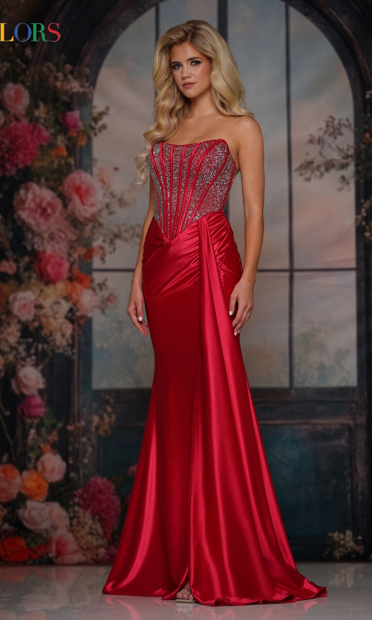 A woman models the Colors Dress 3933, a strapless long corset prom gown featuring an embellished fitted bodice, posing gracefully before a floral arrangement and arched window backdrop.